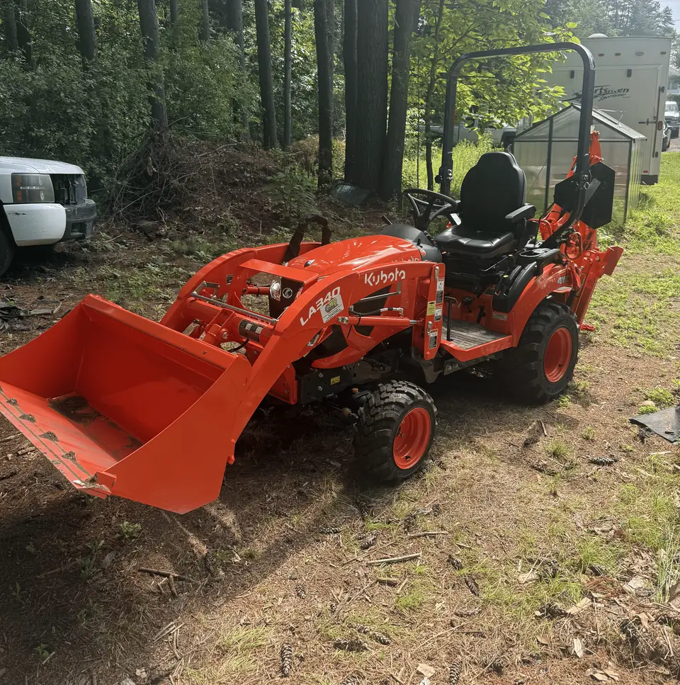 Our compact tractor with loader and backhoe, parked on a dirt lot in front of pine trees
